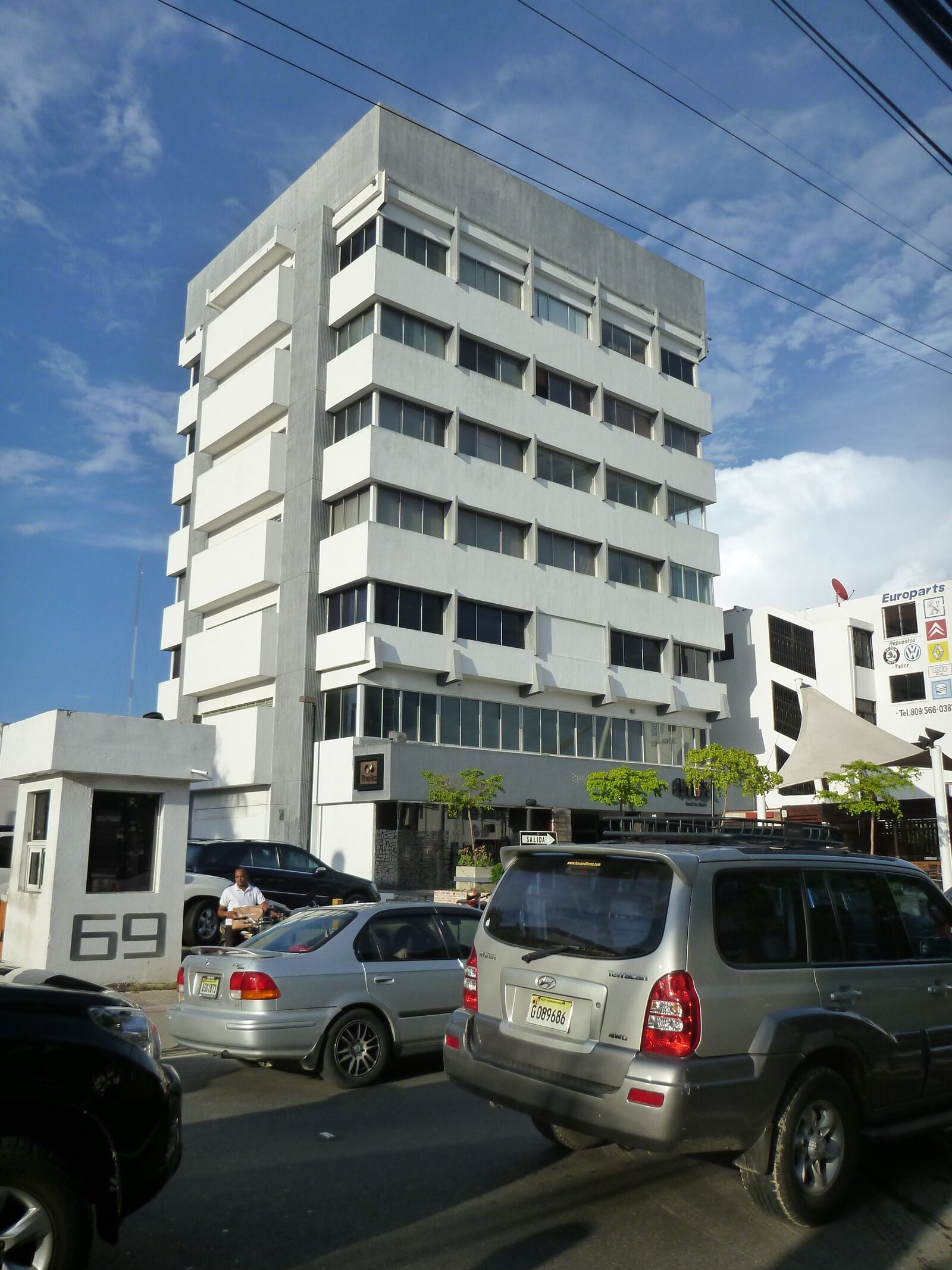 Fotografía del edificio original en la esquina de la calle antes de la renovación, con fachada de concreto blanco y vehículos estacionados al frente.